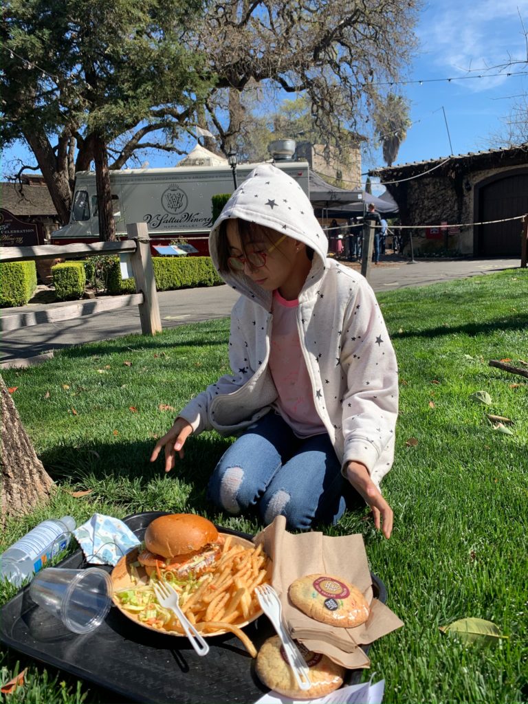 Girl wearing a white hoodie, sitting in the grass eating a burger and french fries at V. Sattui winery in St. Helena, California in Napa Valley