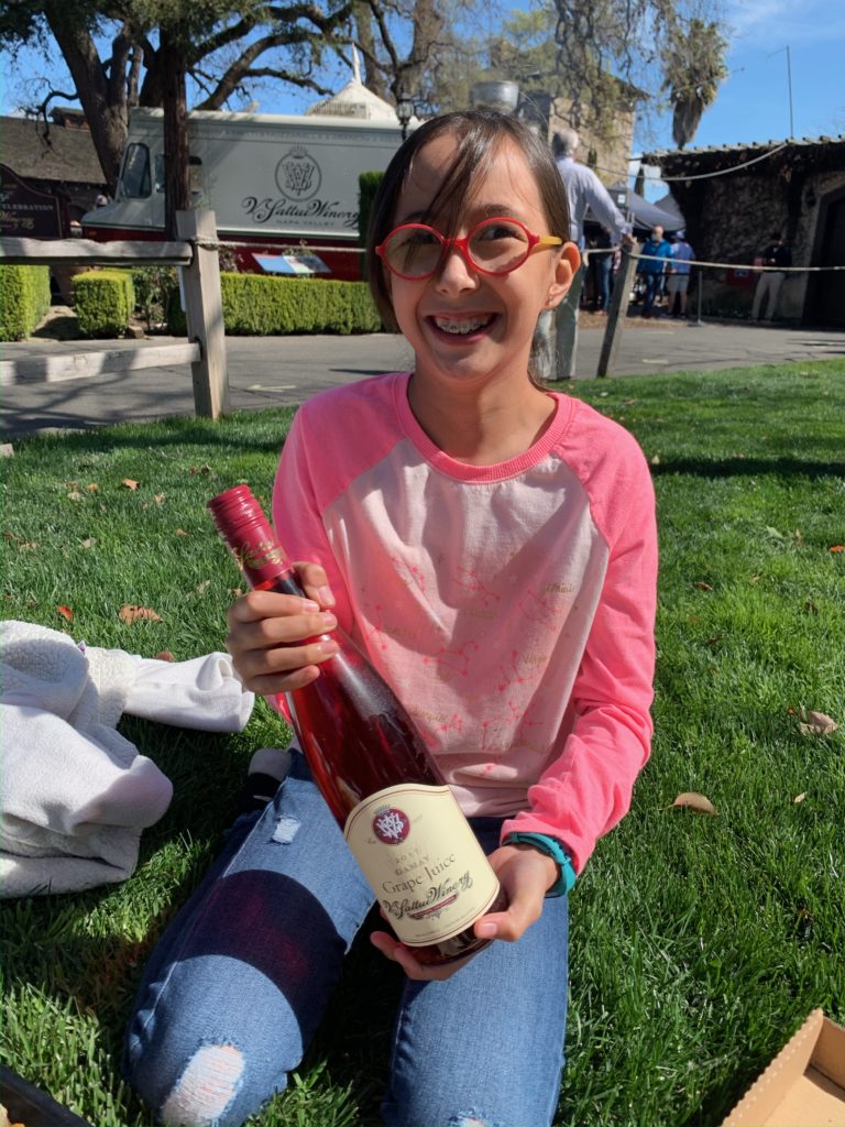 girl wearing red glasses sitting on the grass holding a bottle of grape juice at V. Sattui winery in St. Helena, California in Napa Valley